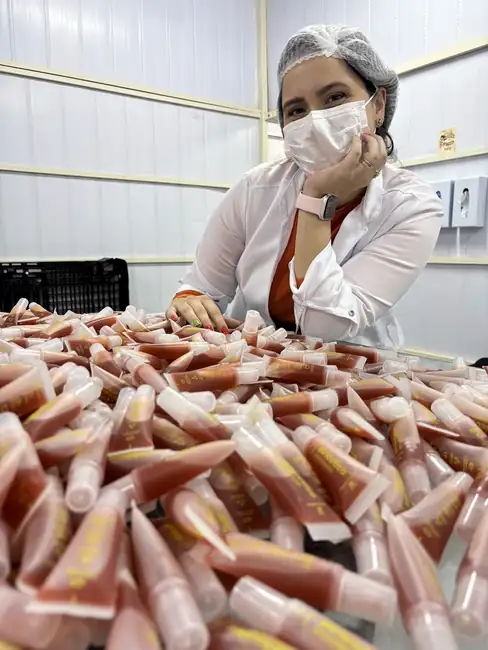 Natali Gutierrez de touca e máscara, apoiada sobre dezenas de bisnagas de gloss na fábrica da Dona Coelha